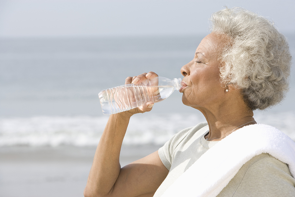 A Black senior woman drinking water from bottle with sea in the background